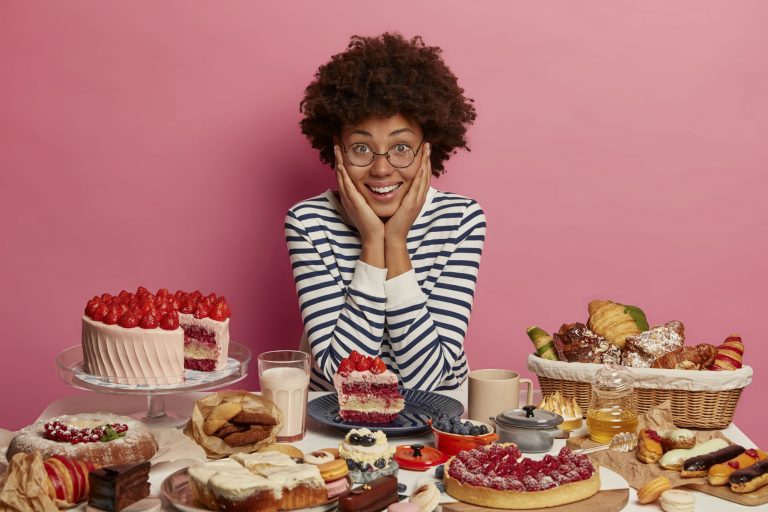 Happy woman behind a table of sugary snacks, including cakes and pies