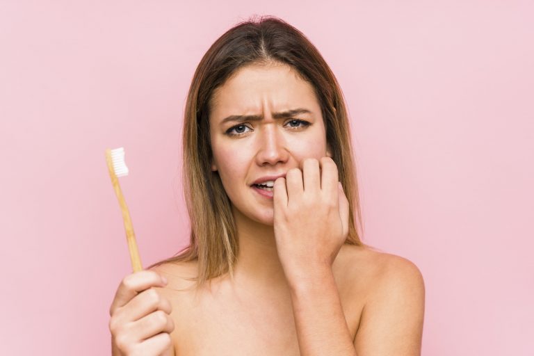 Female with brown hair, appearing stressed, holding a toothbrush in one hand and biting the nails of her other hand