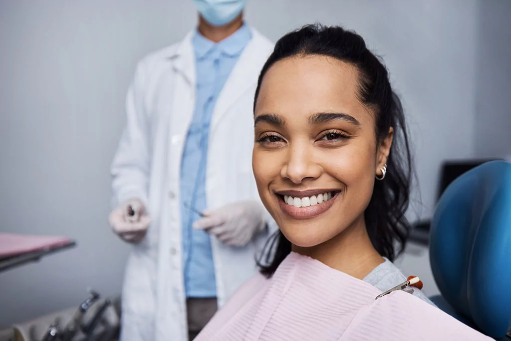 A smiling female patient sitting in a dental chair with a dentist standing in the background.