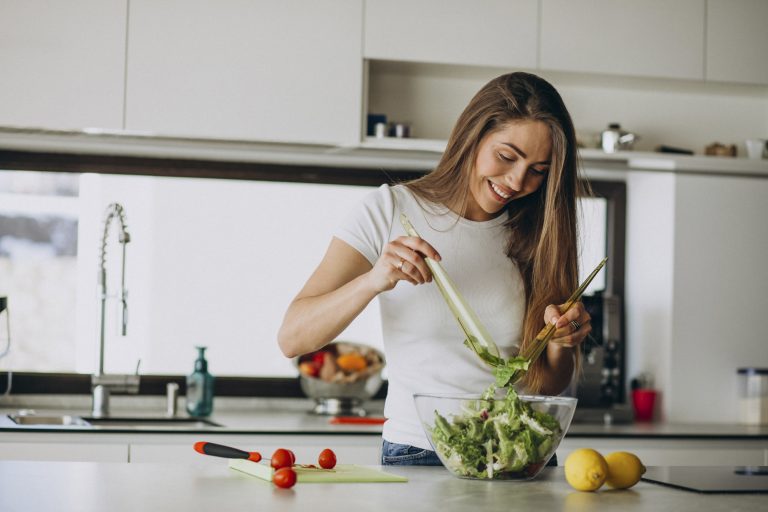 Young female at a kitchen counter, holding two utensils and mixing a salad
