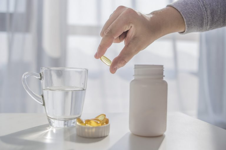 Person holding a nutritional supplement, in front of a table that has a glass of water and other supplements on it